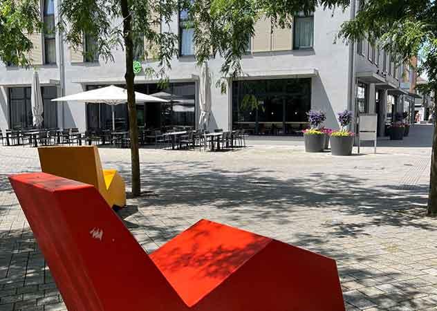 Kornhausplatz Göppingen mit einer roten Bank im Vordergrund unter schattigen Bäumen