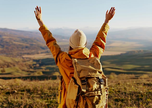 Eine Frau mit Rucksack steht vor einer schönen Naturlandschaft und streckt die Arme in die Luft © Adobestock SHOTPRIME STUDIO