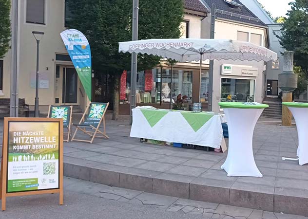 Stand der TEAM KLIMA Oase mit Beachflag, Sonnenschirm, Tischen und einem Aufsteller "Die nächste Hitzewelle kommt bestimmt".