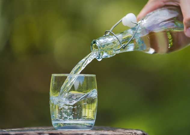 Ein Glas steht auf einem Baumstumpf. Jemand schenkt frisches Wasser aus einer Glasflasch ein ©Adobestock – Naypong-Studio