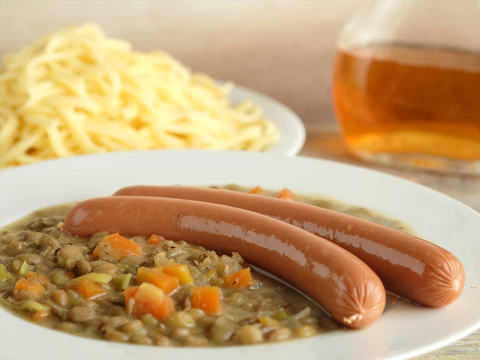 Teller mit Linsen und veganen Würstchen im Hintergrund Spätzle und ein Krug Essig © Adobestock 